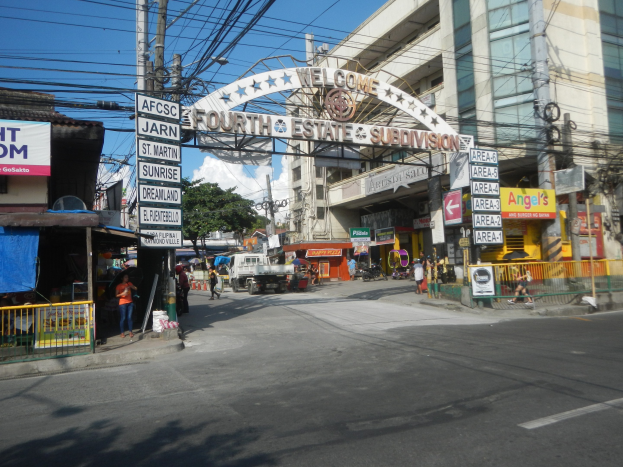 Eine belebte Stadtstraße mit fahrenden Fahrzeugen und Fußgängern, die ein Tor, Gebäude, Strommasten, Bäume und einen bewölkten Himmel zeigt, mit einem "Fourth Estate Subdivision"-Schild in der Mitte der Straße.