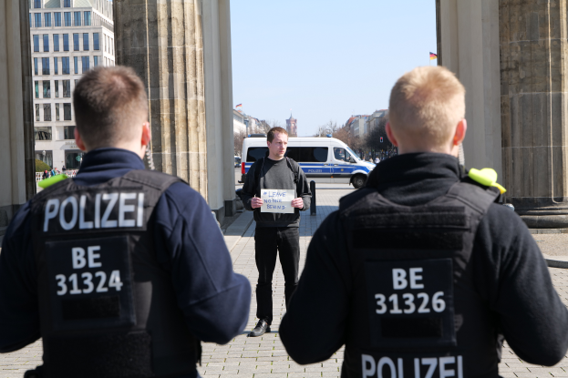 Drei Polizeibeamte stehen vor einem Gebäude, ein Mann in der Mitte hölt ein Blatt Papier, flankiert von Säulen, mit Fahrzeugen, Menschen, Bäumen, Gebäuden und Himmel im Hintergrund.