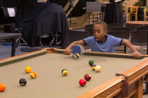 Ein Junge hält einen Tischtennisschläger vor einem Snookertisch mit Billardkugeln, mit Stühlen und anderen Gegenständen im Hintergrund.