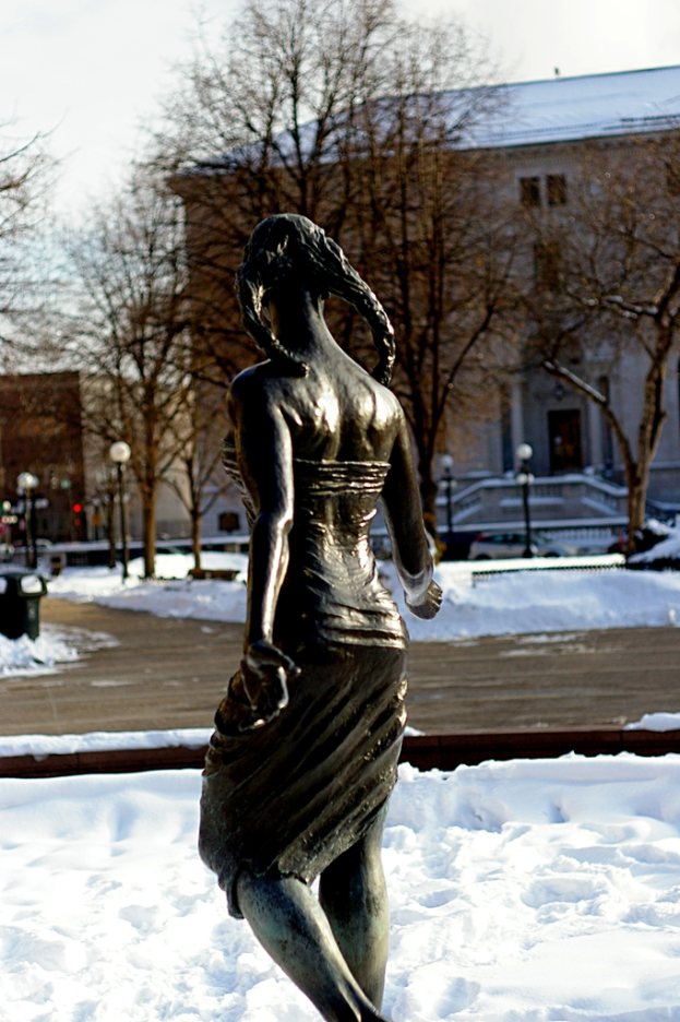Eine Frau-Statue steht im Schnee, mit einem hölzernen Gehweg davor, umgeben von Bäumen, Gebäuden und Laternenpfählen im Hintergrund.