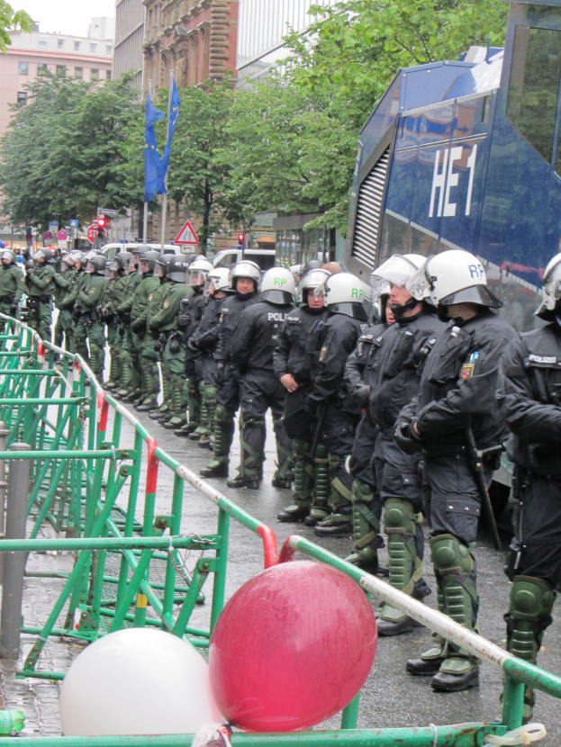 Eine Gruppe von Polizisten in Helmen und mit Waffen vor einer Barriere auf einer Straße mit Geländern und Luftballons, mit Bäumen, Gebäuden, Fahnen und einem klaren blauen Himmel im Hintergrund.