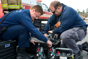 Zwei Männer in blauen Uniformen arbeiten an einem Feuerwehrauto, umgeben von Menschen, Fahrzeugen, Bäumen und einem klaren blauen Himmel, mit verschiedenen Werkzeugen und Geräten in den Händen.