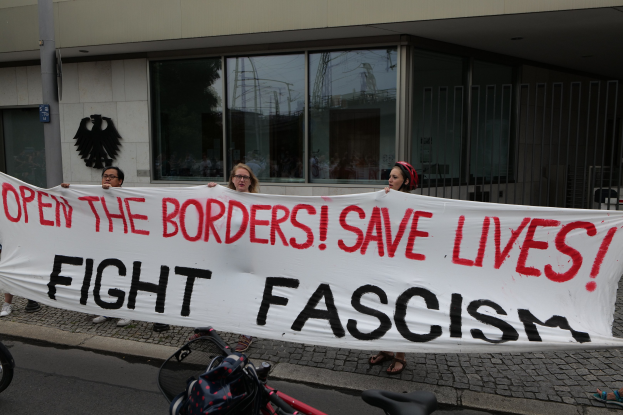 Demonstranten halten ein Transparent mit der Aufschrift "Öffnet die Grenzen, rettet Leben, kämpft gegen Faschismus" vor einem Gebäude mit Glasfenstern und einem Pfahl, mit einem Fahrrad und einer Tasche auf der Straße.