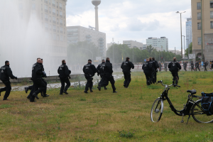 Eine Gruppe von Polizisten, die über ein grünes Feld geht, mit einem Fahrrad im Vordergrund, Gebäuden, Bäumen, Polen, Straßenlaternen, einem Turm und einem klaren blauen Himmel im Hintergrund.