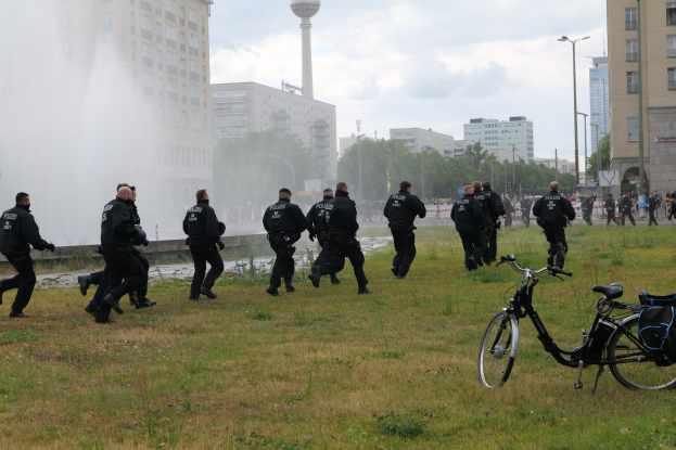 Eine Gruppe von Polizisten, die über ein grünes Feld geht, mit einem Fahrrad im Vordergrund, Gebäuden, Bäumen, Polen, Straßenlaternen, einem Turm und einem klaren blauen Himmel im Hintergrund.