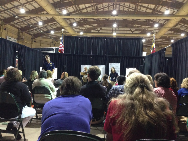 Eine Gruppe von Menschen sitzt auf Stühlen und schaut auf eine Person, die an einem Podium steht, mit Bannern, blauen Vorhängen, Fahnen und Deckenleuchten im Hintergrund.