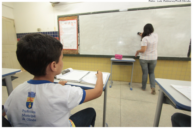 Ein Junge sitzt an einem Tisch und schreibt in ein Buch, während eine Frau die Tafel mit einem Putzlappen putzt in einem Klassenzimmer mit mehreren Tischen, einem haltend eine Kiste, und ein Plakat an der Seite.
