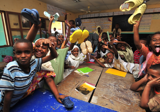 Kinder in einem Klassenzimmer, die fröhlich ihre Schuhe hochhalten, mit Büchern und anderen Gegenständen auf den Tischen, Texttafeln an den Wänden, ein Fenster links und Deckenleuchten und ein Ventilator.