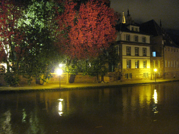 Ein Fluss fließt durch eine Stadt in der Nacht, beleuchtet von Straßenlaternen und gesäumt von Gebäuden mit Fenstern, Bäume säumen die Flussufer unter einem sichtbaren Nachthimmel.