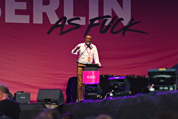Ein Mann in einem weißen Hemd und braunen Hosen steht auf einer Bühne und hält ein Mikrofon in der Hand, mit einem pinken Banner dahinter, das 'Berlin as Fuck' lautet und Lautsprecher auf beiden Seiten.