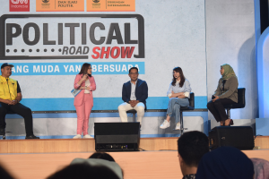 Eine Gruppe von Menschen sitzt auf einer Bühne mit einer Frau, die ein Mikrofon in der Mitte hält, einem Publikum im Vordergrund und einem Banner mit der Aufschrift "Politischer Roadshow 2019" im Hintergrund.