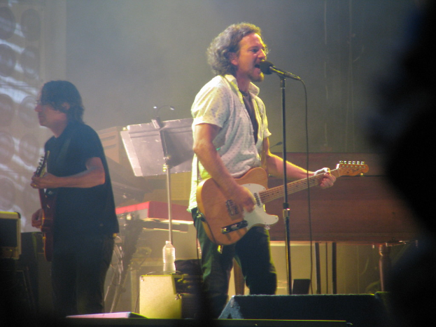 Ein Mann in einem weißen Hemd spielt Gitarre und singt in ein Mikrofon auf der Bühne, mit einem anderen Mann in einem schwarzen Hemd, der Gitarre spielt, und einem Piano, Kabeln, Lautsprechern und Lichtern im Hintergrund.