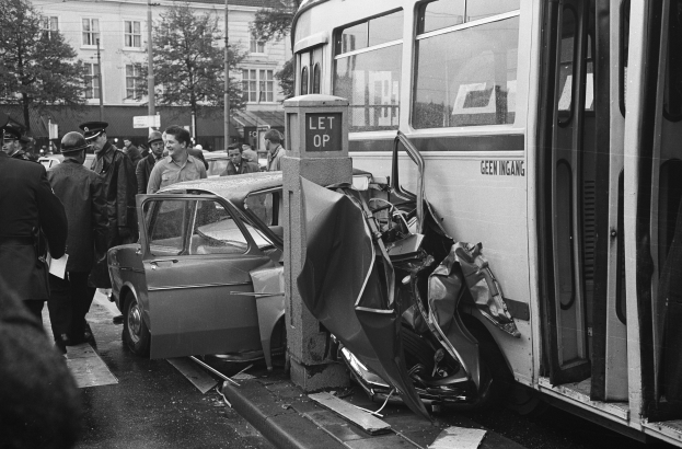 Schwarz-weißes Bild eines Bus, der auf der Seite der Straße in ein Auto gekracht ist, mit Menschen und Gebäuden im Hintergrund.