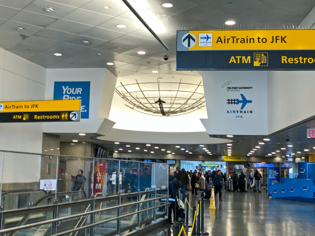 Eine Gruppe von Menschen geht durch eine Flughafenhalle mit einer Rolltreppe auf der linken Seite, Schilder an den Wänden einschließlich einem, das "Airtrain nach JFK" lautet, und Lampen an der Decke.