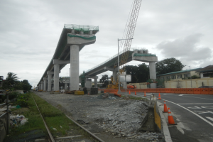 Eine Baustelle mit einer Brücke im Hintergrund, eine Straße mit Absperrgittern auf der rechten Seite, Steine und Gras auf dem Boden, eine Eisenbahnschiene auf der linken Seite, Bäume und Gebäude auf beiden Seiten der Straße und ein bewölkter Himmel.