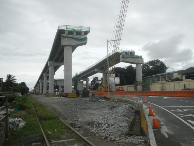 Eine Baustelle mit einer Brücke im Hintergrund, eine Straße mit Absperrgittern auf der rechten Seite, Steine und Gras auf dem Boden, eine Eisenbahnschiene auf der linken Seite, Bäume und Gebäude auf beiden Seiten der Straße und ein bewölkter Himmel.