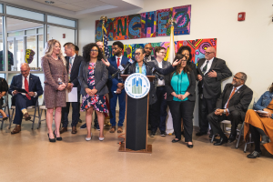 Gruppe von Menschen vor einem Rednerpult mit Mikrofon und einer Tafel mit der Aufschrift "Sancho-Stadtrat tritt dem Stadtrat bei", mit sitzenden und stehenden Teilnehmern, Flaggen, einem Feuerlöscher und Deckenleuchten im Hintergrund.