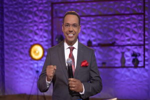 Stephen Colbert, der einen Anzug, Krawatte und eine Brille trägt, steht auf einer Bühne und hält ein Mikrofon in der Hand, mit einer Wand und verschiedenen Gegenständen dahinter.