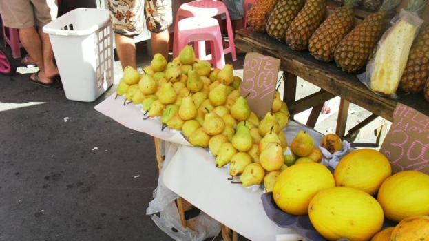 Eine Marktszene mit verschiedenen Fruächten auf einem Gestell, ein paar Höcker, einige Menschen und zwei Preiszettel.