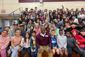 Gruppe von Schülern, die in einem Turnsaal für ein Foto posieren, einige sitzen auf der Treppe und andere stehen, eine Wand im Hintergrund, eine Tür und ein Ausgangsschild links.
