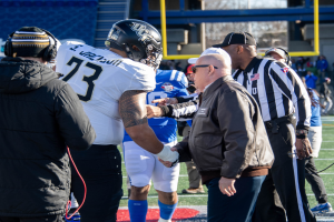 Ein Fußballspieler mit Helm gibt einem Schiedsrichter die Hand auf dem Feld, umgeben von einer Gruppe von Menschen, mit einem Zaun, Pfosten, einem Gebäude, Bäumen und dem Himmel im Hintergrund.