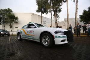 Ein weißer Polizeiwagen steht vor einem Gebäude in Mexiko-Stadt, umgeben von einer Gruppe Menschen in verschiedenenfarbigen Kleidern und Mützen, mit Bäumen und einem klaren blauen Himmel im Hintergrund.