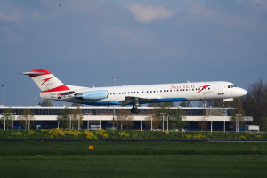 Ein Austrian-Airlines-Airbus-A320-200-Flugzeug startet am Frankfurter Flughafen, mit grünem Gras, blühenden Pflanzen, Bäumen, Laternenmasten und einem Gebäude im Hintergrund unter einem bewölkten Himmel.