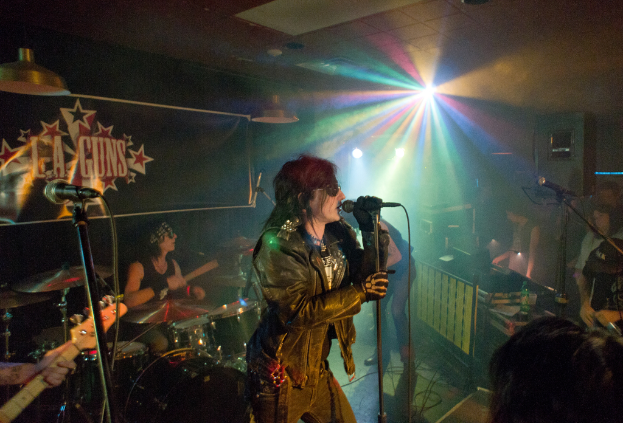 Eine Frau in einer schwarzen Jacke singt auf der Bühne in ein Mikrofon, während ein Mann mit Sonnenbrille und Stirnbinde hinter ihr ein Instrument spielt und ein Plakat mit "LA Guns" im Hintergrund zu sehen ist.