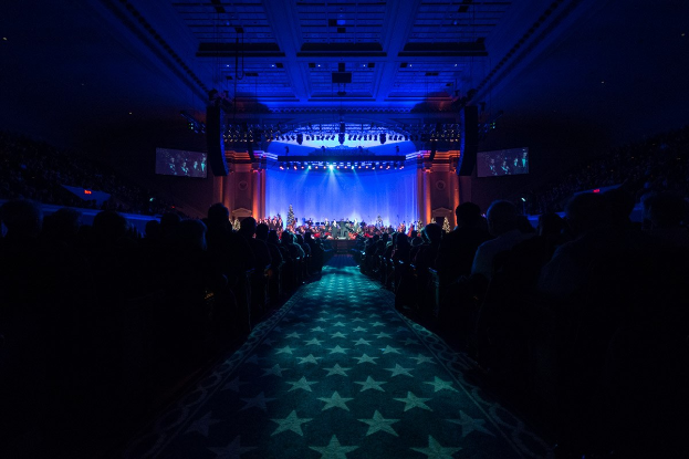 Ein großer Saal mit Reihen von Stühlen, die auf eine Bühne zeigen, auf der Musiker spielen, unter einer Decke mit Lichtern und Fernsehern, was auf eine Konzerthalle hinweist.