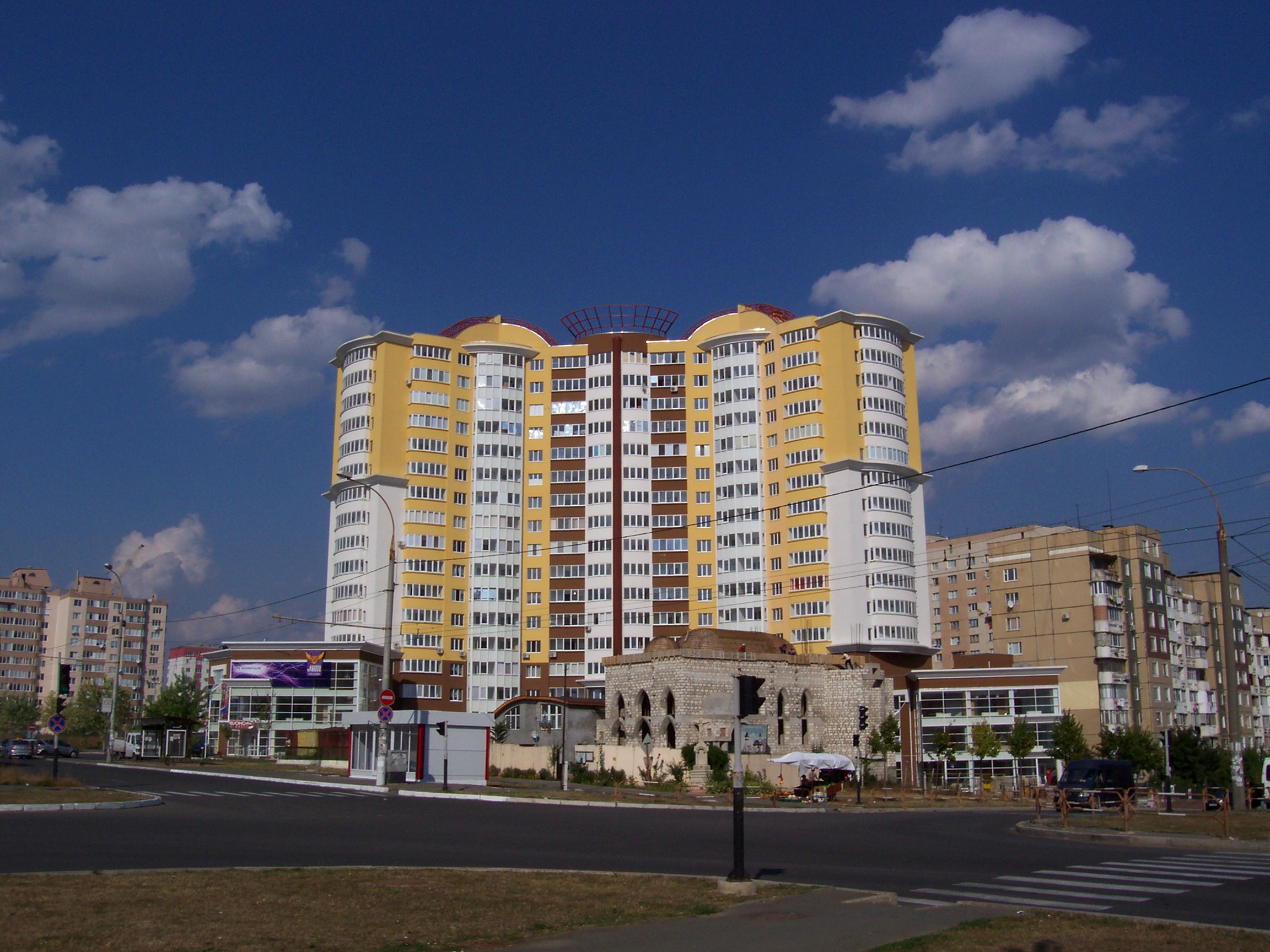 Großes Wohnhaus in städtischer Umgebung mit Straßeninfrastruktur, Fahrzeugen, Bäumen und bewölktem Himmel.