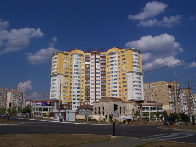 Großes Wohnhaus in städtischer Umgebung mit Straßeninfrastruktur, Fahrzeugen, Bäumen und bewölktem Himmel.