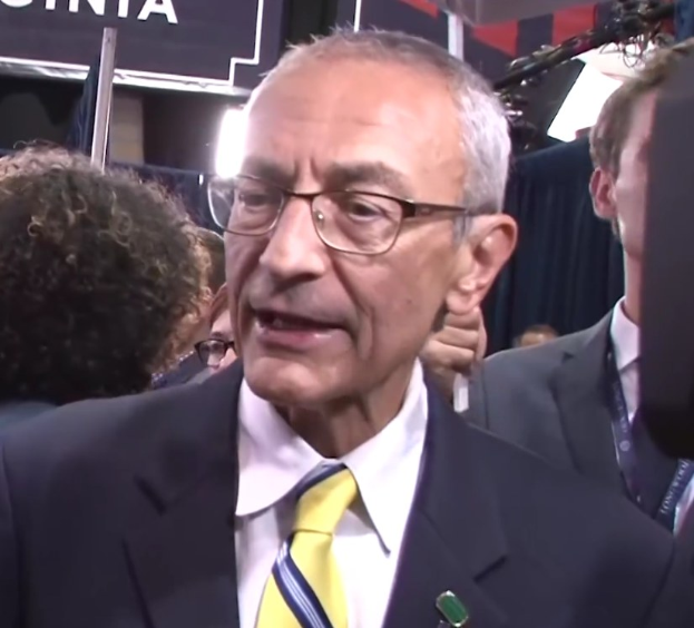 Ein Mann im Anzug und Krawatte spricht zu Reportern, trägt eine Brille und steht hinter einem Mikrofon mit Stativ, umgeben von einer Gruppe von Menschen, mit Lampen und einer Tafel mit Schrift im Hintergrund.