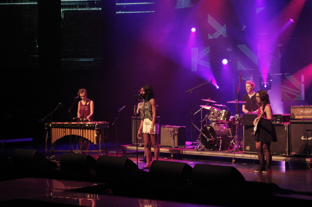 Vier Personen, die auf einer Bühne mit Musikinstrumenten und einem Mikrofon auftreten.