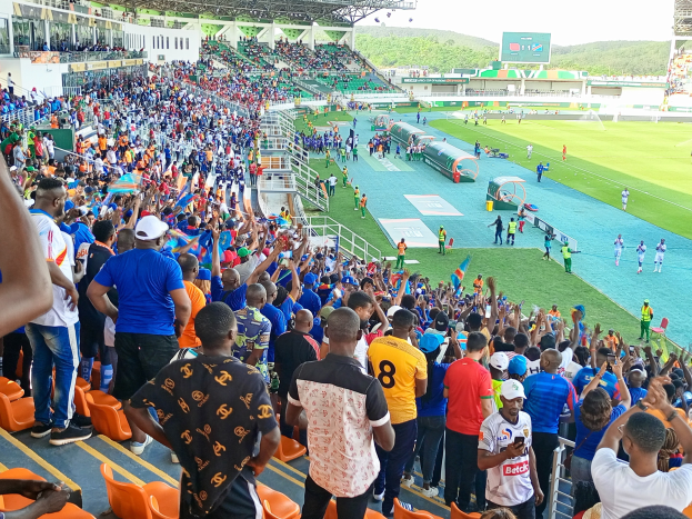 Eine große Menschenmenge versammelt sich vor einem eingezäunten Fußballfeld mit Hügeln und einem klaren blauen Himmel und wartet gespannt auf den Beginn eines Spiels während der Afrikameisterschaft 2019.