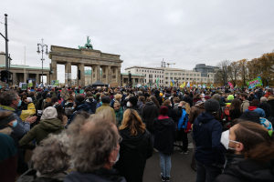 Eine große Menschenmenge steht vor dem Brandenburger Tor in Berlin, Deutschland, viele tragen Mützen und Masken, einige halten Schilder, mit Laternen, Bäumen, Gebäuden mit Fenstern und Bögen und einer Statue auf einem Pfeiler im Hintergrund unter einem bewölkten Himmel.