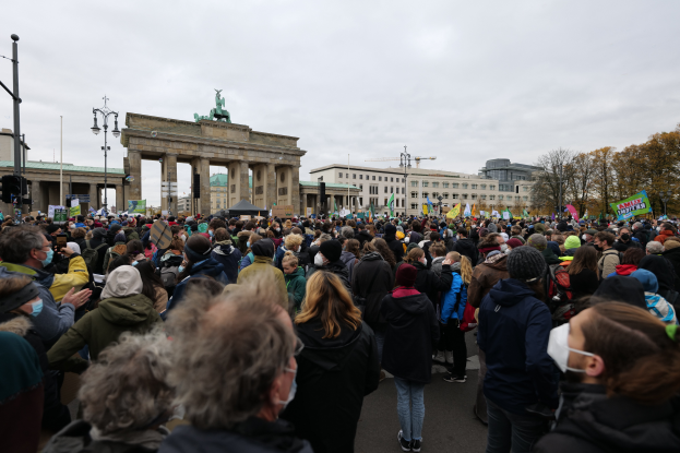 Eine große Menschenmenge steht vor dem Brandenburger Tor in Berlin, Deutschland, viele tragen Mützen und Masken, einige halten Schilder, mit Laternen, Bäumen, Gebäuden mit Fenstern und Bögen und einer Statue auf einem Pfeiler im Hintergrund unter einem bewölkten Himmel.