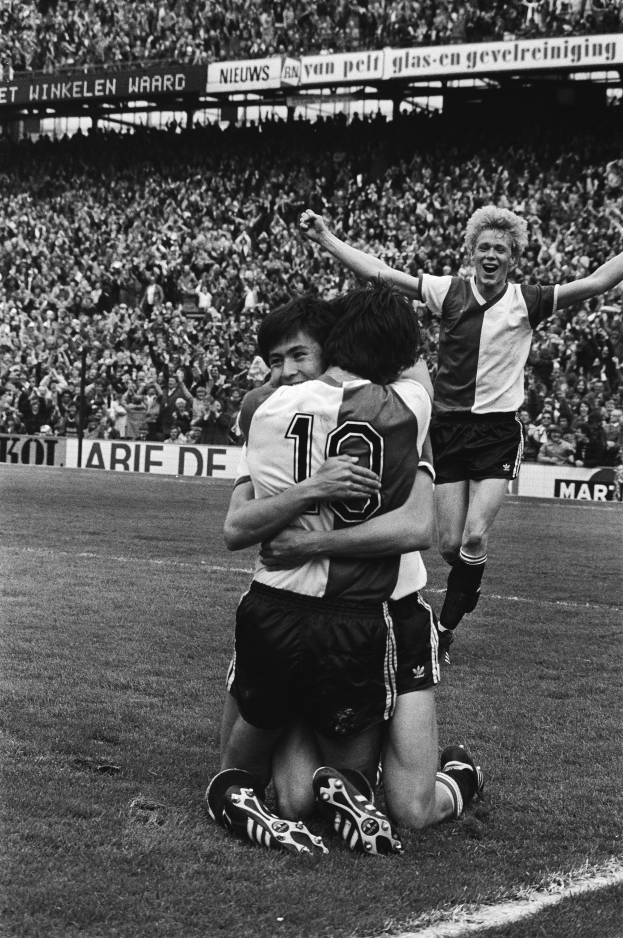 Drei Fussballspieler umarmen sich auf dem Boden und feiern ein Tor, mit einer jubelnden Menge und schwenkenden Fahnen im Hintergrund, dargestellt in Schwarz-Weiss.