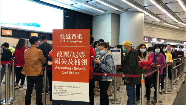 Eine Gruppe von Menschen mit Masken steht in einer Schlange an einem Flughafen, umgeben von Absperrpollern mit Bändchen, mit einer Tafel im Vordergrund, auf der "Hong Kong Airport Refunds Due to Covid-19" steht, und Pfeilern und Deckenlampen im Hintergrund.
