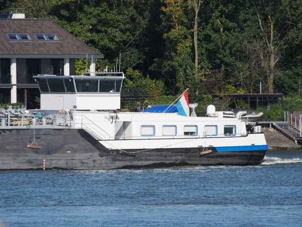 Ein großes Fährenboot fährt einen Fluss hinunter, mit einem Haus auf der linken Seite und Bäumen im Hintergrund.