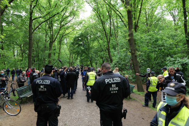Eine Gruppe von Polizeibeamten steht vor einer Menschenmenge, mit Fahrrädern und einer Bank im Vordergrund, Bäume und Himmel im Hintergrund.