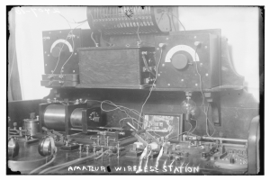 Ein altes Schwarz-Weiß-Foto einer Amateur-Funkstation mit verschiedenen elektronischen Geräten und Drähten, die miteinander verbunden sind, und Text am unteren Rand.