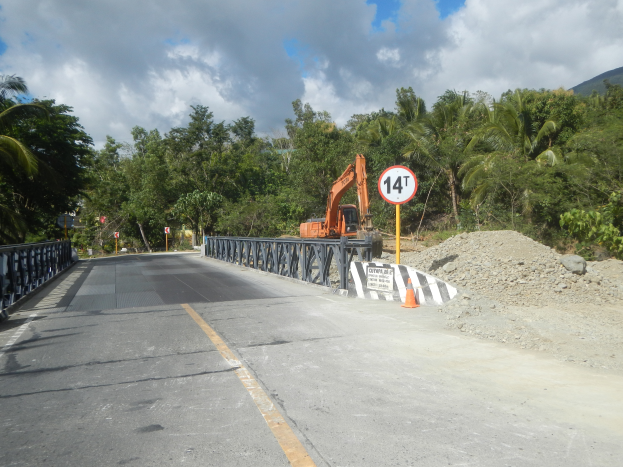 Eine Brücke mit Geländern, ein Schild an einem Pfahl, ein Bagger, Verkehrsabsperrungen, Steine, Bäume, Hügel und ein bewölkter Himmel, mit einer Baustelle und einem Kran auf der Seite der Straße.