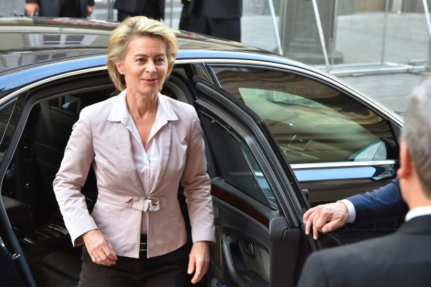 Ursula von der Leyen, eine Frau in einer rosafarbenen Jacke und schwarzen Hosen, steigt aus einem Auto aus, neben ihr steht ein Mann im Anzug, während einige Menschen auf der Straße und ein Zaun im Hintergrund zu sehen sind.
