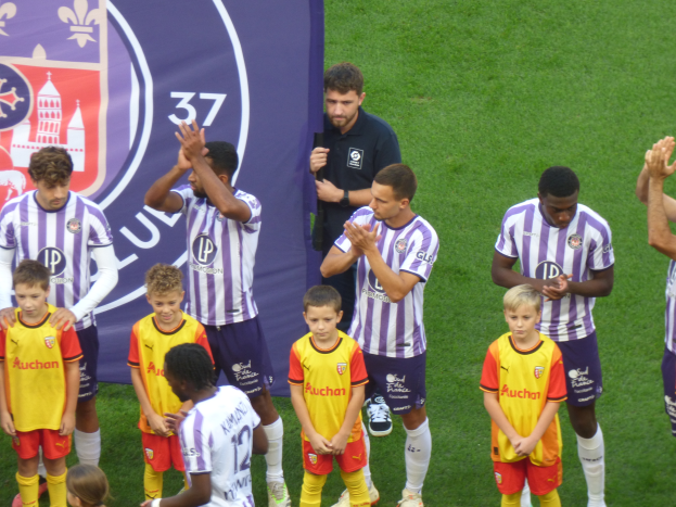 Eine Gruppe junger Jungs steht auf einem gr├╝nen Fussballfeld und klatscht in die H├Ąnde, mit einem Banner und Logos im Hintergrund.