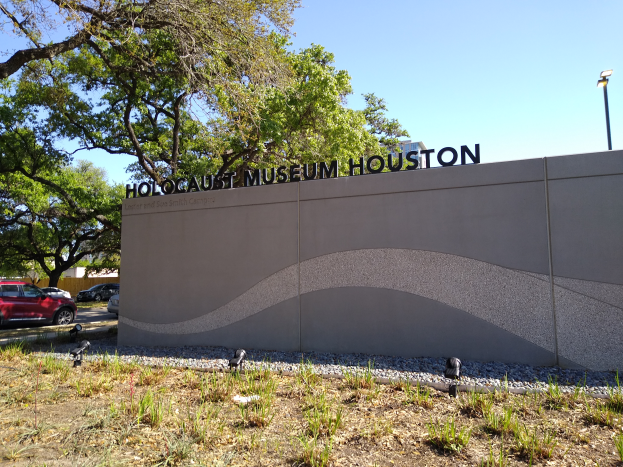 Das Holocaust-Museum in Houston, Texas, mit einer textbedeckten Wand, Steinen, Gras, Pflanzen, Bäumen, Fahrzeugen auf der Straße, einem Straßenschild und einem bewölkten Himmel.