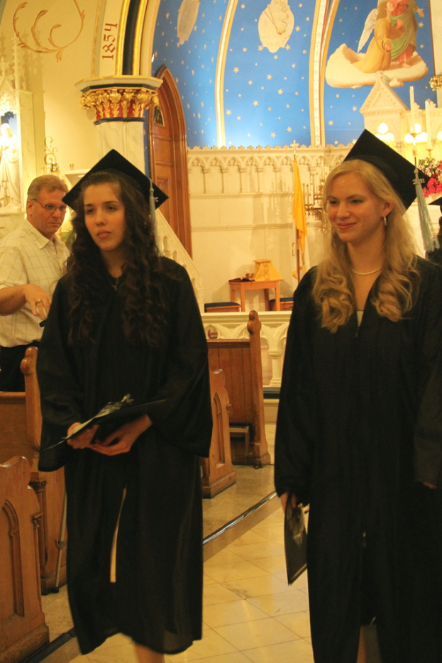 Zwei Frauen, die Papiere oder Zertifikate halten, wobei eine Frau lächelt; ein Mann steht neben Bänken; ein Tisch mit Hockern, eine gelbe Flagge, eine Treppe, eine Säule und eine Tür im Hintergrund; eine weiße Wand mit einem blauen Abschnitt oben.