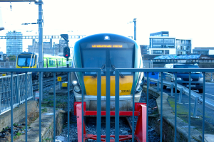 Zwei Züge auf dem Gleis mit einem Zaun im Vordergrund, Masten auf dem Land, einer mit einer Signallampe, ein Auto auf der Straße rechts, Gebäude im Hintergrund und der Himmel oben.
