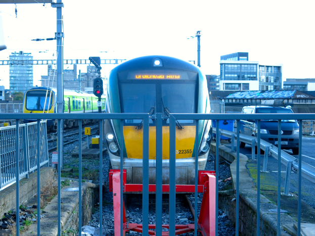 Zwei Züge auf dem Gleis mit einem Zaun im Vordergrund, Masten auf dem Land, einer mit einer Signallampe, ein Auto auf der Straße rechts, Gebäude im Hintergrund und der Himmel oben.