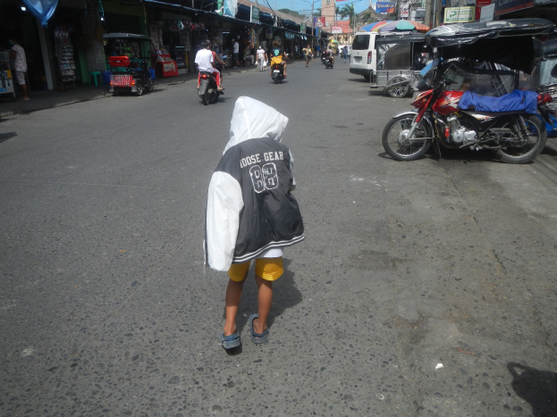 Ein junger Junge geht eine Straße entlang, die von Motorrädern und anderen Fahrzeugen bevölkert ist, mit Gebäuden, Namensschildern, Bäumen, Polen und einem klaren blauen Himmel im Hintergrund.