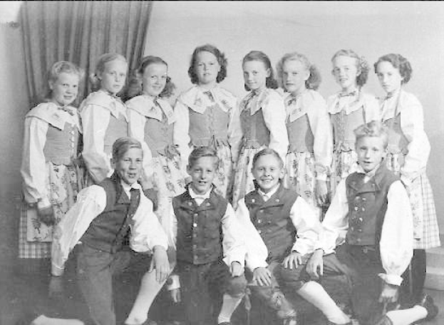 Schwarzes und weißes Foto einer Gruppe von Kindern, die vor einer Wand mit einem Vorhang posieren, mit der Beschriftung "1930er Kinder-Schule" unten.